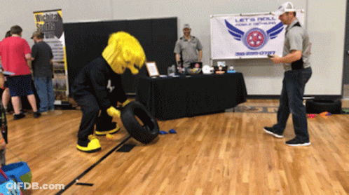 A Mascot Is Rolling A Tire In Front Of A Sign That Says Let 'S Roll GIF