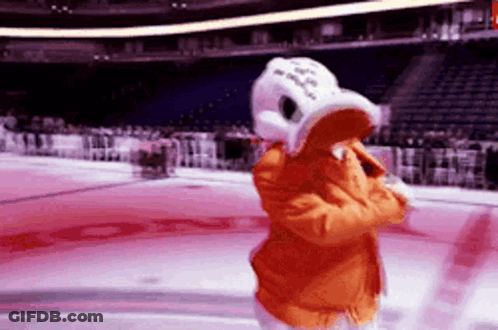 A Mascot Is Standing On A Hockey Rink Wearing An Orange Jacket And A White Hat GIF