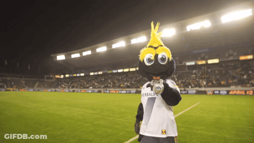 A Mascot Wearing A Shirt That Says Herbalife Stands On A Soccer Field GIF