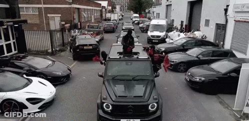 A Mercedes Benz G63 Is Driving Down A Street Surrounded By Lots Of Cars . GIF