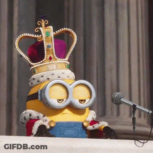 A Minion Wearing A Crown And Goggles Is Sitting At A Podium In Front Of A Microphone GIF