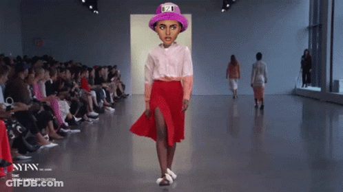 A Model Walks Down A Runway Wearing A Pink Hat And A Red Skirt GIF