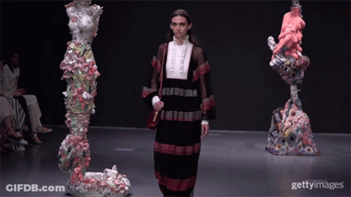 A Model Walks Down The Runway At A Fashion Show In Front Of Sculptures GIF