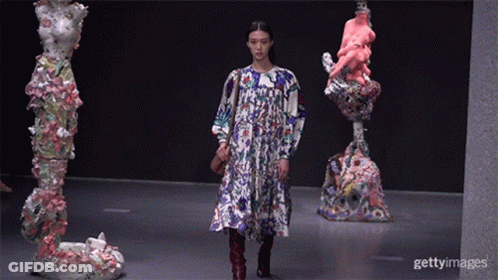 A Model Walks Down The Runway At A Fashion Show In Front Of Statues GIF