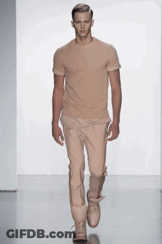 A Model Walks Down The Runway At A Fashion Show Wearing A Tan T-Shirt And Tan Pants GIF
