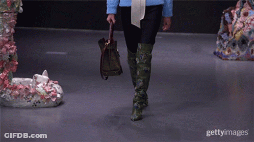 A Model Walks Down The Runway At A Fashion Show While Holding A Purse And Wearing Green Boots GIF