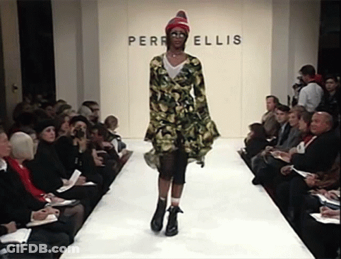 A Model Walks Down The Runway In Front Of A Wall That Says Perri Ellis GIF