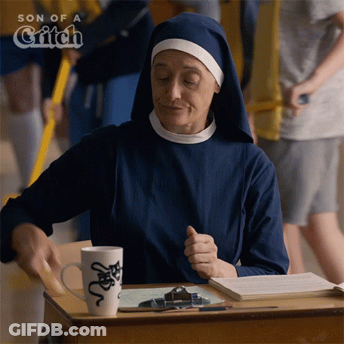A Nun Sits At A Desk In Front Of A Sign That Says Son Of A Witch GIF