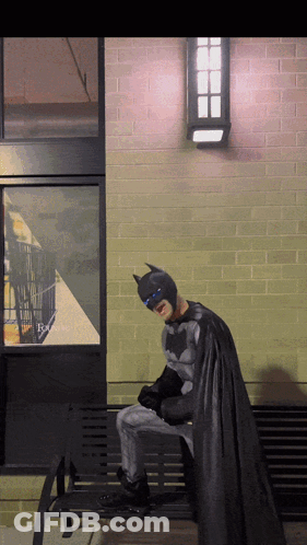 A Person Dressed As Batman Sits On A Bench GIF