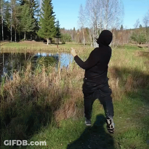 A Person In A Black Hoodie Is Standing In A Field Near A Lake GIF