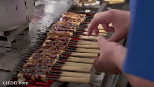 A Person In A Blue Shirt Is Cooking A Row Of Skewers On A Grill GIF