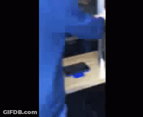 A Person In A Blue Suit Is Standing Next To A Wooden Table With A Cell Phone On It . GIF