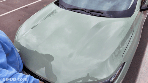 A Person In A Blue Sweatshirt Is Looking Under The Hood Of A Green Car GIF