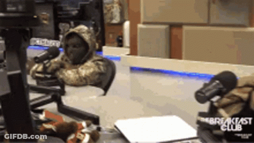 A Person In A Hooded Jacket Is Sitting In Front Of A Breakfast Club Microphone GIF