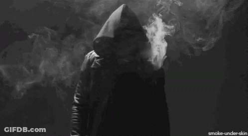 A Person In A Hooded Jacket Is Smoking A Cigarette In A Dark Room GIF