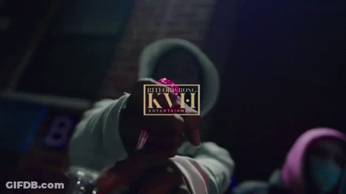 A Person In A Hoodie Holds A Red Object In Front Of A Sign That Says Kvh GIF