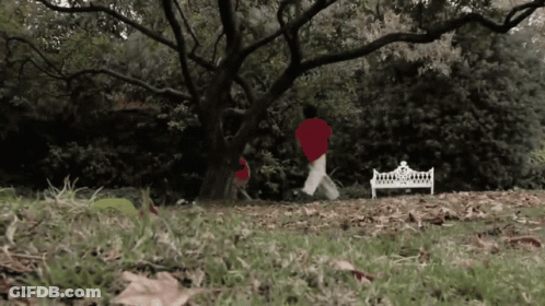A Person In A Red Shirt Is Running In A Park Near A White Bench GIF