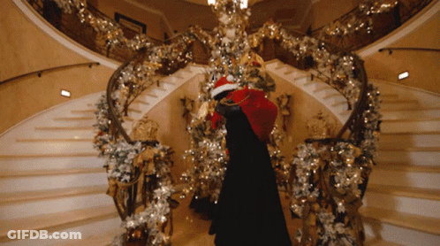 A Person In A Santa Hat Is Standing On A Staircase With Christmas Decorations GIF