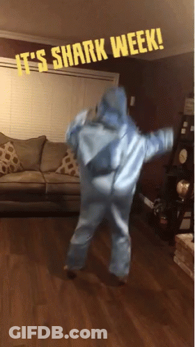 A Person In A Shark Costume Is Dancing In A Living Room With A Sign That Says It 'S Shark Week GIF