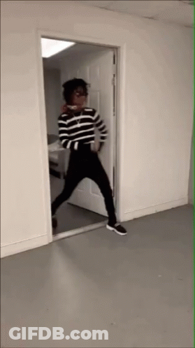 A Person In A Striped Shirt And Black Pants Stands In A Doorway GIF