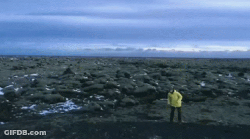 A Person In A Yellow Jacket Stands On A Rocky Beach GIF