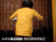A Person In A Yellow Shirt Is Standing In Front Of A Curtain And Says `` Good Morning '' . GIF