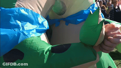 A Person In An Inflatable Costume With A Blue Cape Holds Another Person 'S Hand GIF
