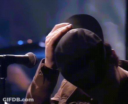 A Person Is Covering Their Face With A Hat While Standing In Front Of A Microphone GIF