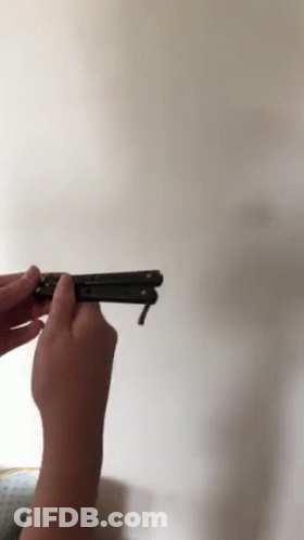 A Person Is Holding A Black Butterfly Knife In Front Of A White Wall GIF