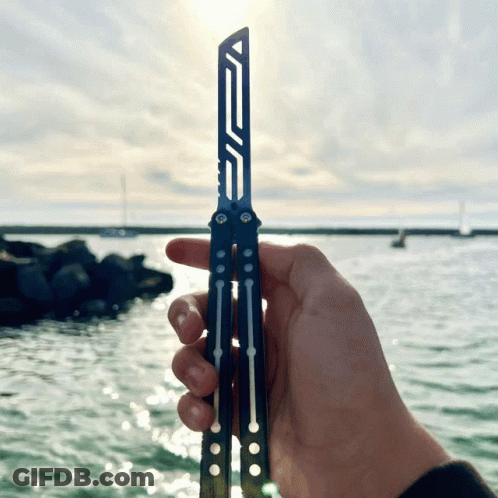 A Person Is Holding A Butterfly Knife In Front Of The Water GIF