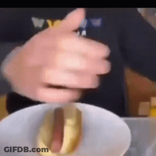 A Person Is Holding A Hot Dog With Mustard On A Plate . GIF