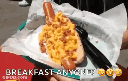 A Person Is Holding A Hot Dog With Scrambled Eggs On It And The Words Breakfast Anyone Below It GIF