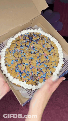 A Person Is Holding A Pizza In A Box That Looks Like A Cookie Cake GIF