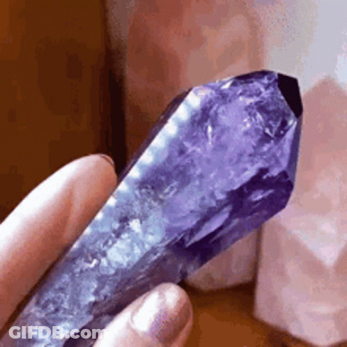 A Person Is Holding A Purple Crystal In Their Hand In Front Of Some Pink Crystals . GIF