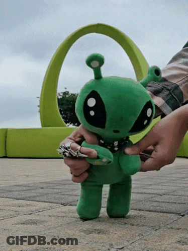A Person Is Holding A Stuffed Green Alien In Front Of A Green Arch GIF