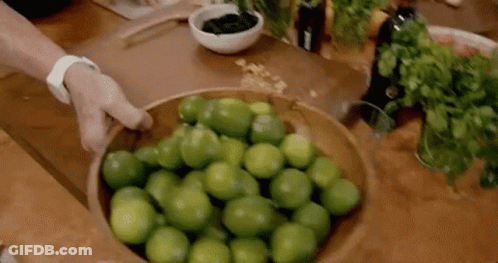 A Person Is Holding A Wooden Bowl Filled With Limes On A Table . GIF