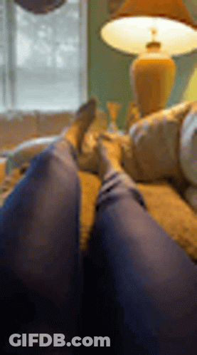 A Person Is Laying On A Couch With Their Feet Up In Front Of A Lamp . GIF