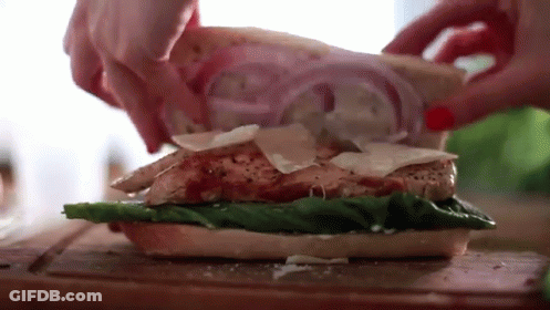 A Person Is Making A Sandwich With Chicken , Lettuce , Cheese And Onions On A Cutting Board . GIF