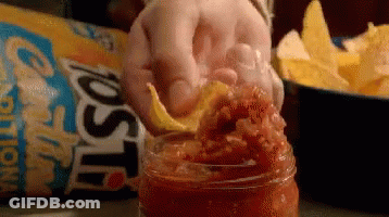 A Person Is Putting A Tortilla Chip Into A Jar Of Salsa . GIF