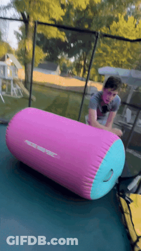 A Person Is Rolling On A Pink And Blue Inflatable Cylinder On A Trampoline GIF