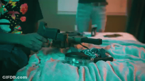 A Person Is Sitting On A Bed Holding A Gun And A Lighter . GIF