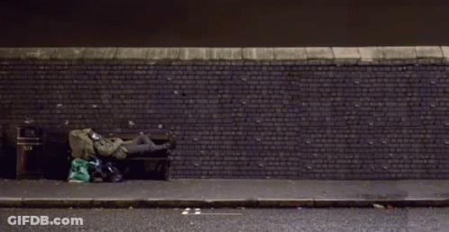 A Person Is Sleeping On A Bench In Front Of A Brick Wall With A Trash Can On The Sidewalk GIF