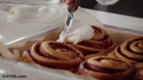A Person Is Spreading Frosting On Cinnamon Rolls GIF