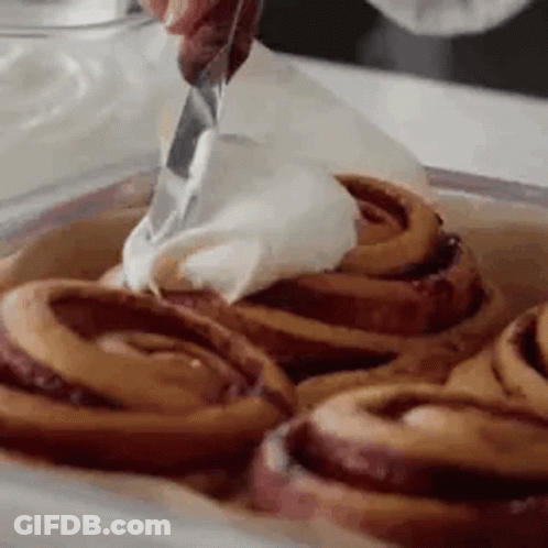 A Person Is Spreading Frosting On Cinnamon Rolls In A Pan . GIF