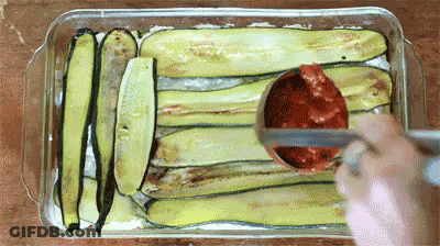 A Person Is Spreading Tomato Sauce On A Casserole Dish Filled With Zucchini Slices . GIF