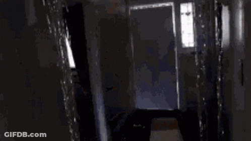 A Person Is Standing In A Dark Hallway Looking Out A Doorway . GIF