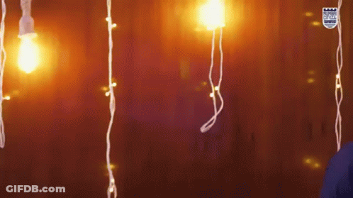 A Person Is Standing In Front Of A Wall With Christmas Lights Hanging From It GIF