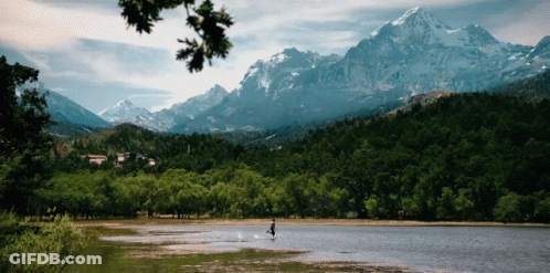 A Person Is Standing In The Middle Of A Lake With Snowy Mountains In The Background GIF
