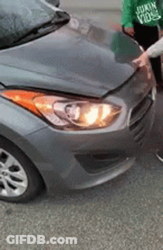 A Person Is Standing Next To A Gray Car With A Broken Hood And Headlight . GIF