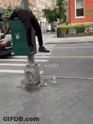 A Person Is Standing On A Green Trash Can On A Street GIF